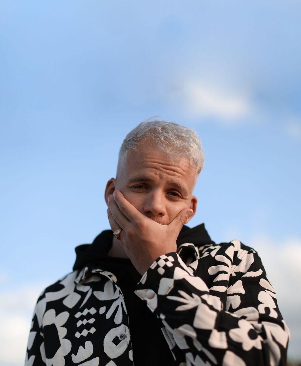 Portrait de l'artiste avec le ciel en fond, il se couvre le bas du visage avec la main.