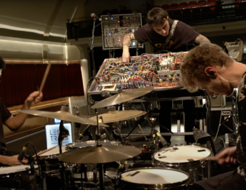 Deux artistes jouent de la batterie face à face, un troisième manipule les boutons et les câbles d'une grande console sonore. Ils sont en studio.