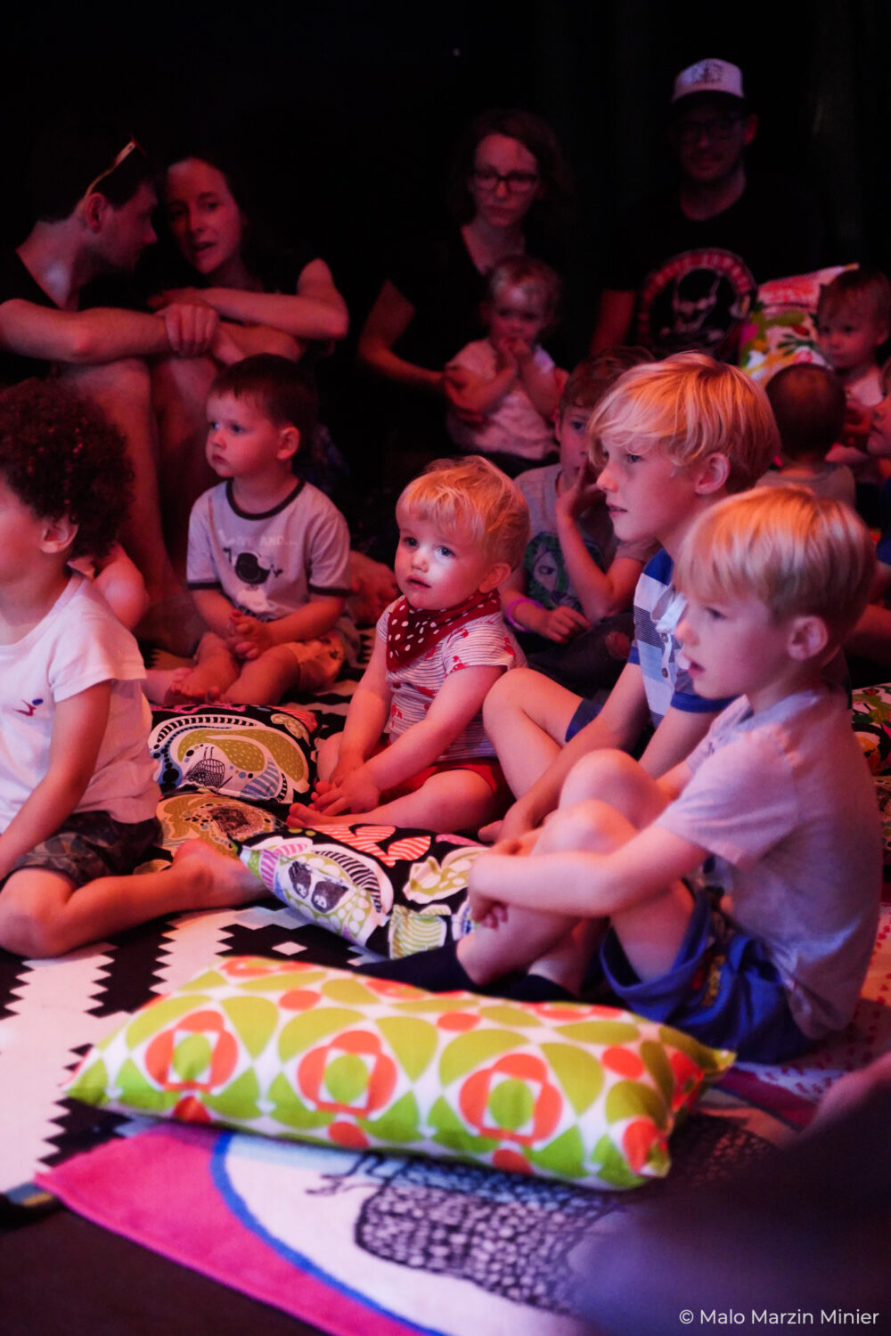 Le public est un groupe d'enfants assis sur des tapis et coussins au sol