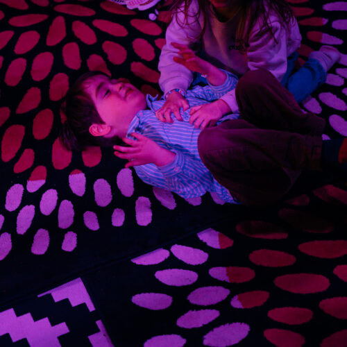 Un enfant allongé sur un tapis se fait chatouiller par une autre enfant.