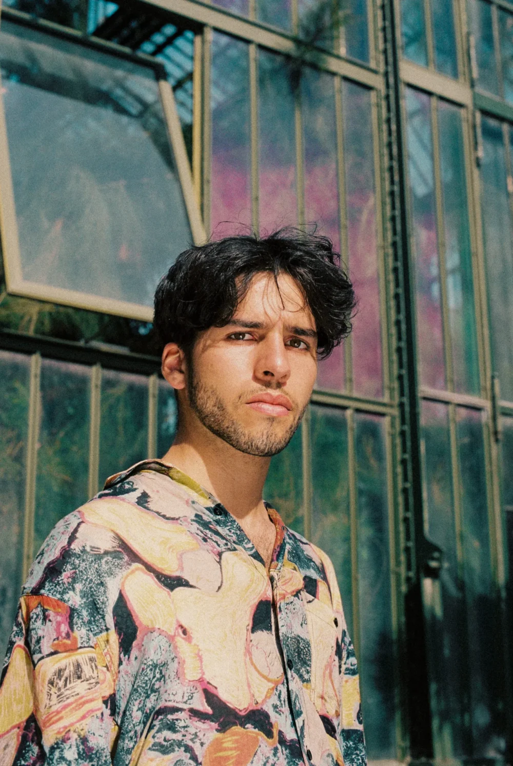 L'artiste pose avec une chemise bariolée devant un bâtiment de verre. Il a le regard pénétrant.