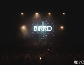 Vue d'ensemble d'un concert depuis le fond de salle, assez sombre, avec le logo BIRRD très éclairé sur scène