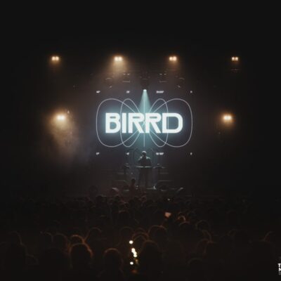 Vue d'ensemble d'un concert depuis le fond de salle, assez sombre, avec le logo BIRRD très éclairé sur scène