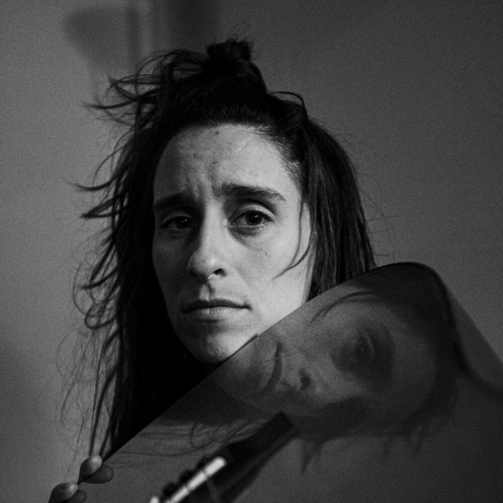 Portrait en noir et blanc d'une femme décoiffée au regard triste dont on voit le reflet dans le bois verni d'une guitare qu'elle tient sous son visage