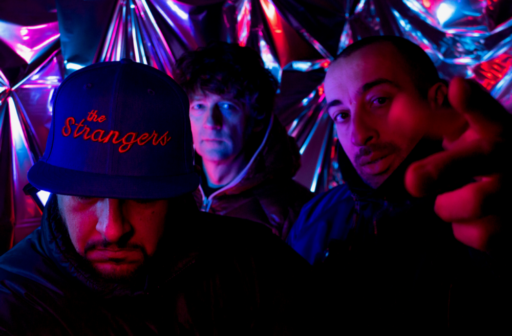 Portrait de trois hommes dans un décor illuminé de lumières de club bleues et rouges. L'un deux baisse la tête pour montrer l'inscription de sa casquette "The Strangers"