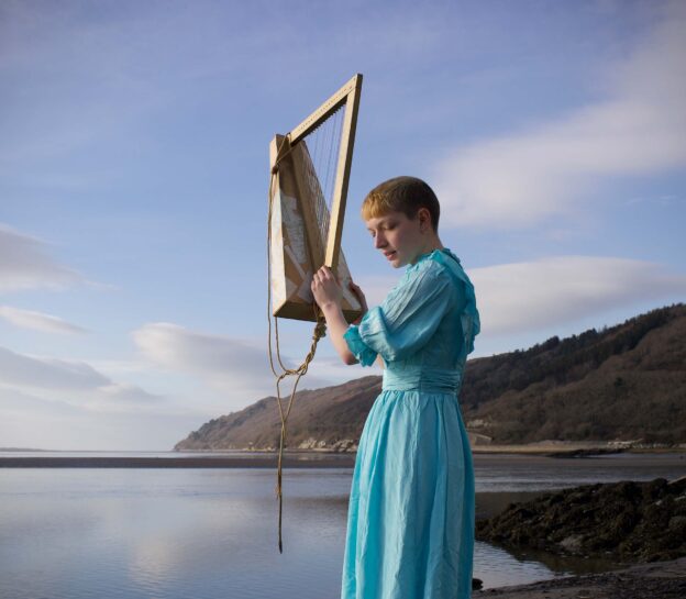 Photo d'une femme manipulant une sorte de harpe D.I.Y avec la mer, le ciel et une île en arrière plan