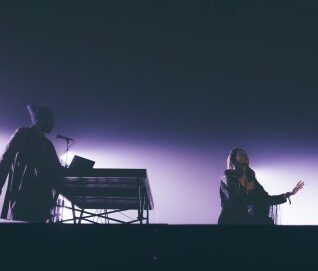 photo de scène sombre avec une lumière violette, une femme chante et un musicien est sur ordinateur