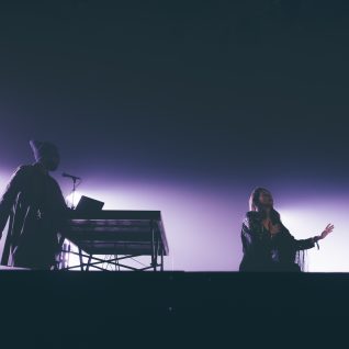 photo de scène sombre avec une lumière violette, une femme chante et un musicien est sur ordinateur