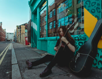 Dans la rue, une jeune femme guitariste joue assise au sol appuyée contre une vitrine colorée