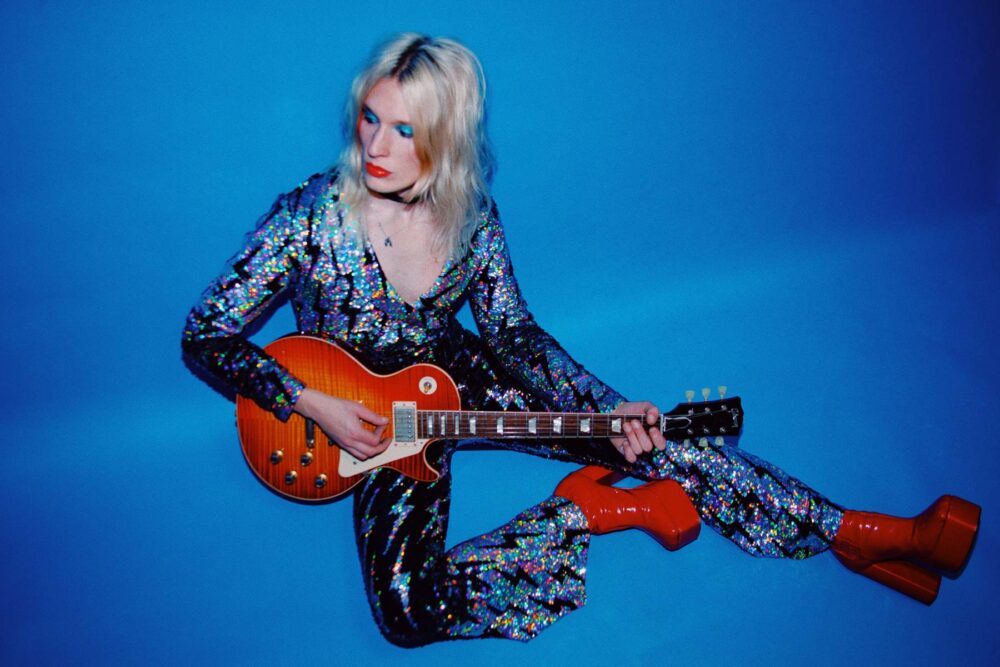 Artiste androgyne avec une combinaison à paillettes jouant de la guitare électrique assis au sol