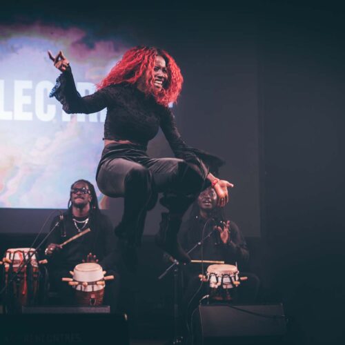 une artiste aux cheveux rouges saute devant deux joueurs de percussions