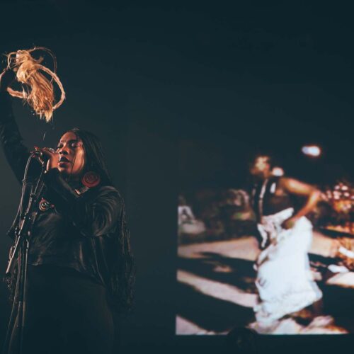 Une artiste chante avec le main en l'air tenant une sorte de fouet de crin tressé