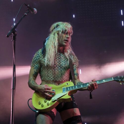 un guitariste masculin habillé en short, bas résilles, haut a strass et perruque blonde